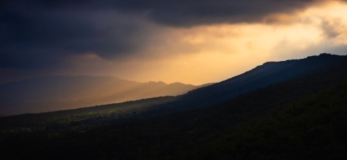 A dramatic landscape at dusk, with dark clouds and golden light illuminating the mountains and valleys below.
