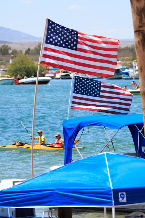 Two American flags wave above a blue boat, with kayakers paddling in the water nearby.