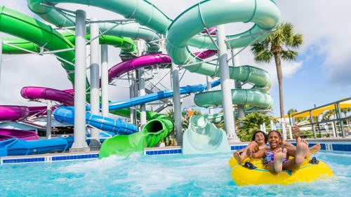 Three children joyfully ride a yellow inflatable tube down a water slide at a colorful water park.