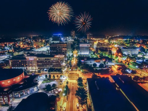 Aerial view of a city at night with colorful fireworks above and illuminated buildings below.