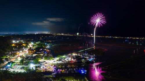 A vibrant night scene of a waterfront city with colorful fireworks illuminating the sky above.