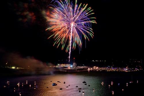 Colorful fireworks burst over a dark sky, illuminating a waterfront with boats and distant lights.