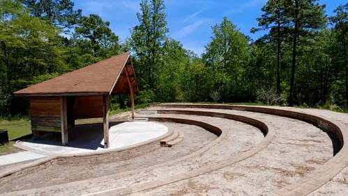 An outdoor amphitheater with curved seating and a wooden stage, surrounded by trees under a clear blue sky.