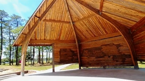 A wooden pavilion with a high, arched ceiling, surrounded by trees and open space.
