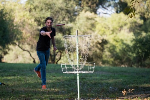 A person throws a disc towards a metal disc golf basket in a green park setting.