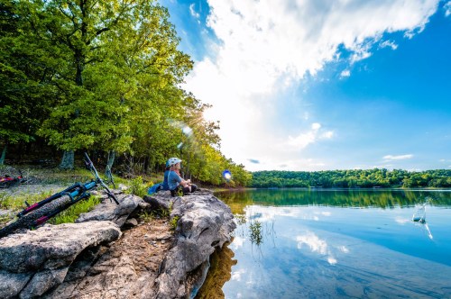 A person sits on a rocky shore by a calm lake, surrounded by trees and blue skies, with a bike nearby.