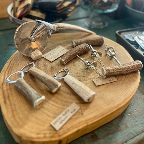 A wooden serving board displays various kitchen tools with antler handles, including a pizza cutter and bottle openers.