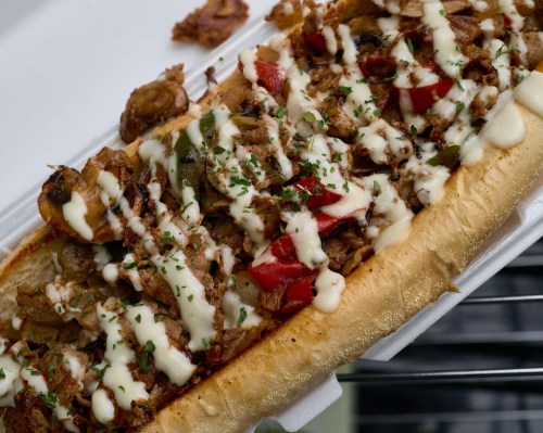 A loaded sandwich with grilled meat, peppers, and drizzled sauce on a toasted hoagie roll, garnished with herbs.
