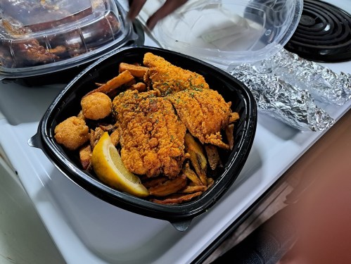 A takeout container with fried fish, seasoned fries, hushpuppies, and a lemon wedge on a kitchen counter.