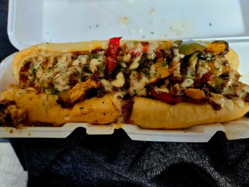 A loaded sandwich with grilled chicken, peppers, and drizzled sauce, served in a white takeout container.