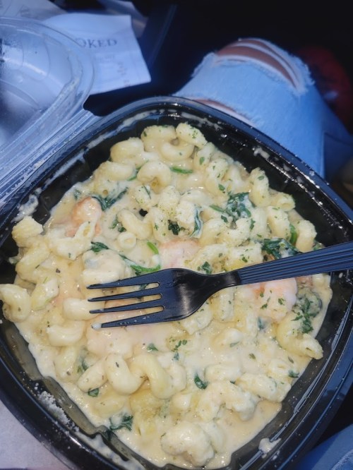 A takeout container filled with creamy macaroni and cheese, garnished with herbs, and a fork resting on top.