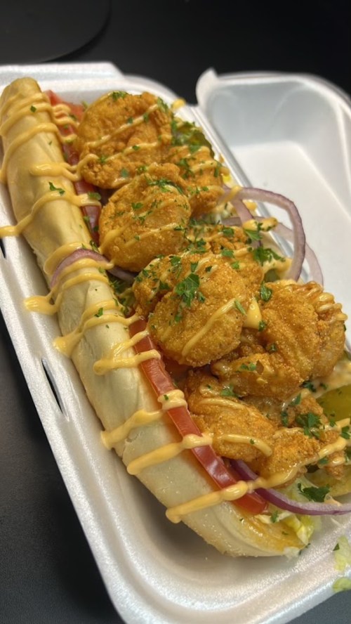 A loaded hot dog topped with crispy shrimp, lettuce, tomato, onions, and drizzled with sauce, served in a foam container.