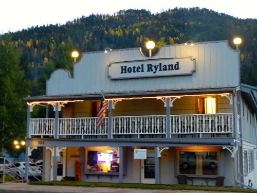 A charming two-story hotel with a sign reading "Hotel Ryland," surrounded by trees and mountains at dusk.