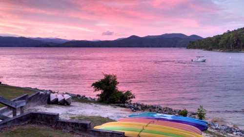 A serene lake at sunset, with colorful kayaks on the shore and a boat cruising on the water against a pink sky.