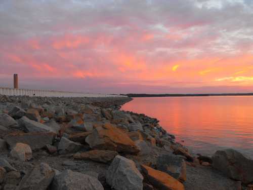 A serene waterfront scene at sunset, with colorful clouds reflecting on calm water and rocky shoreline.