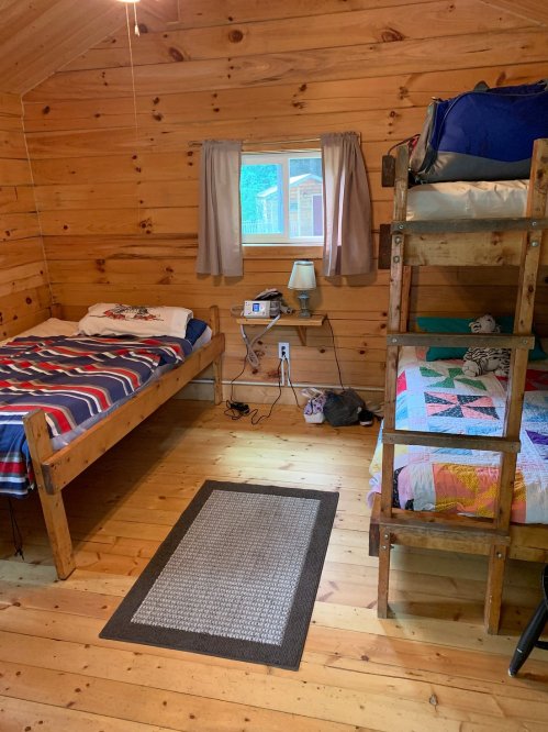 Cozy cabin interior with wooden walls, two beds, a quilt, and a window letting in natural light.