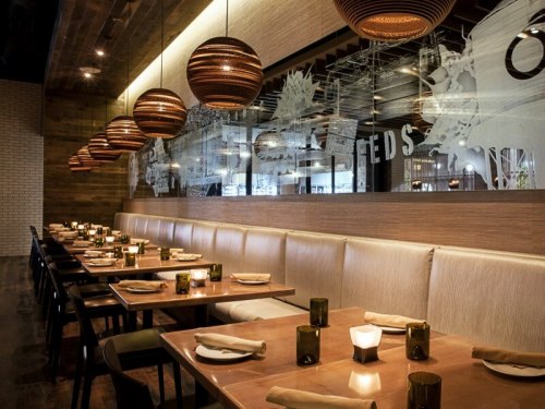 Modern restaurant interior with wooden accents, pendant lights, and a long table set for dining. Soft lighting creates a cozy atmosphere.
