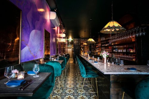 A stylish restaurant interior featuring a bar, green velvet chairs, and purple walls with decorative lighting.