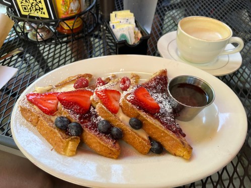 A plate of French toast topped with strawberries, blueberries, and syrup, served with a cup of coffee on a table.