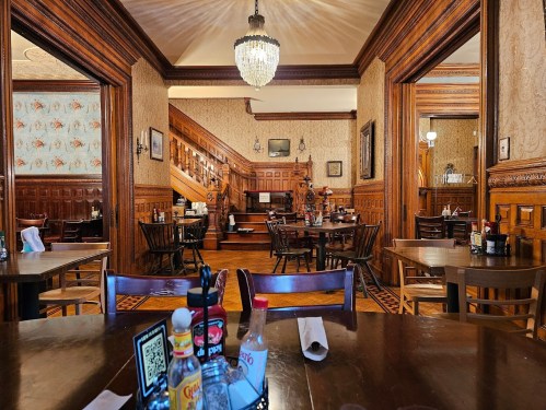 A cozy restaurant interior with wooden decor, tables set for dining, and a staircase in the background.