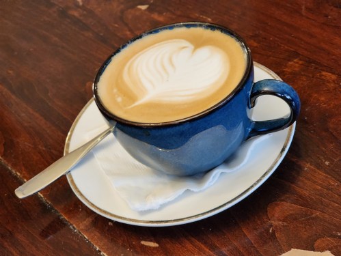 A blue coffee cup with a heart-shaped latte art sits on a white saucer on a wooden table. A spoon rests beside it.