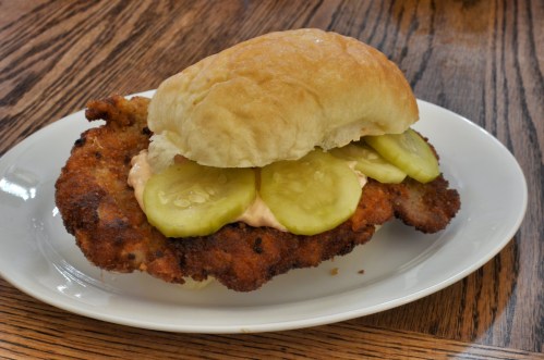 A crispy fried chicken sandwich with pickles and sauce on a soft bun, served on a white plate.