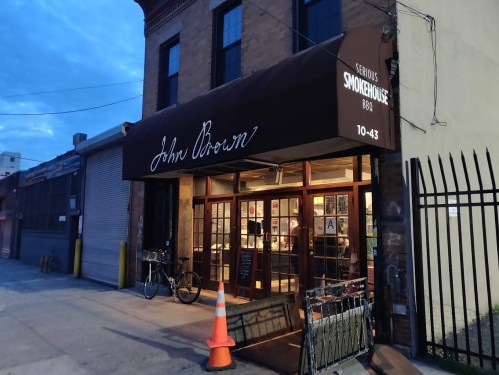 Exterior of John Brown Smokehouse BBQ, featuring a brown awning and large windows, with a bike parked outside.