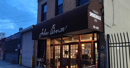 Exterior of John Brown Smokehouse BBQ, featuring a brown awning and large windows, in a dimly lit urban setting.