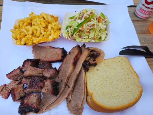 A plate with sliced brisket, macaroni and cheese, coleslaw, and a slice of bread on a white paper surface.