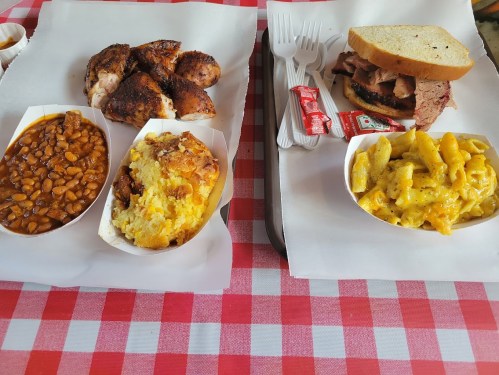 A picnic-style meal featuring grilled chicken, baked beans, cornbread, macaroni and cheese, and a sandwich with meat.
