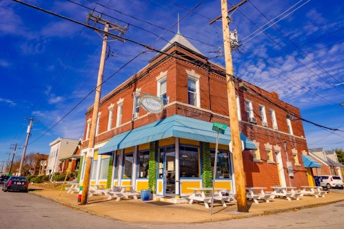 A brick building with a blue awning, outdoor seating, and power lines, set against a clear blue sky.