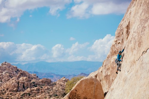 A climber ascends a rocky cliff in a desert landscape under a blue sky with fluffy clouds.