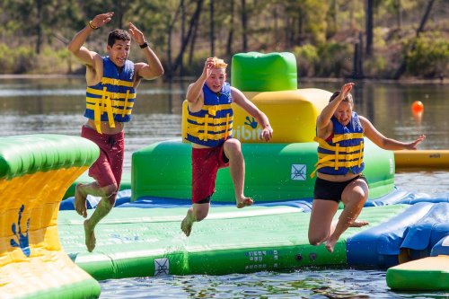 Three people in life jackets jumping off a colorful inflatable water structure into a lake on a sunny day.