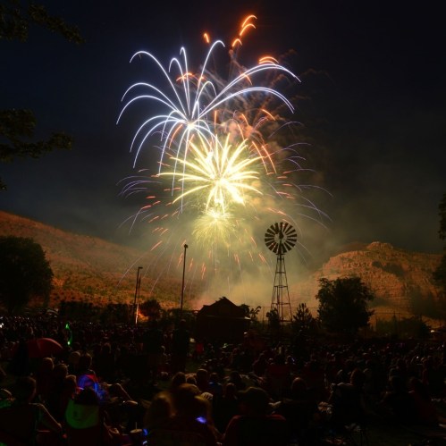 Colorful fireworks illuminate the night sky over a crowd gathered near a windmill and mountains.