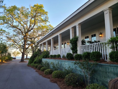 A charming house with a porch, surrounded by greenery and trees, overlooking a serene body of water in the distance.
