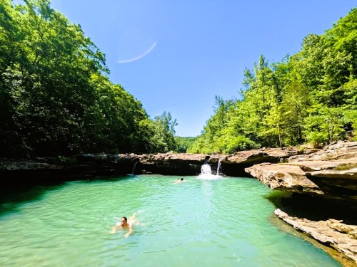 A serene natural pool surrounded by lush green trees, with a small waterfall and two people swimming.