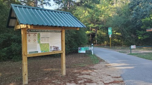 Sign for Kleb Woods Nature Preserve & Center, with a path leading into a wooded area. Green trees in the background.
