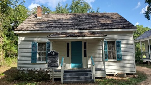A small white house with blue shutters and a front porch, surrounded by trees and a historical marker in front.
