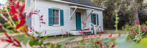 A quaint white house with blue shutters and a front porch, surrounded by greenery and red flowers.
