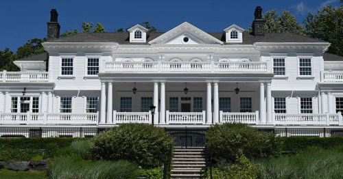 A large white mansion with a grand entrance, multiple columns, and lush greenery in front under a clear blue sky.