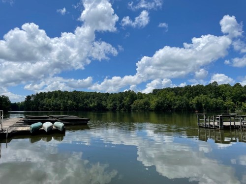 A serene lake scene with boats, wooden docks, and fluffy clouds reflecting on the water under a bright blue sky.