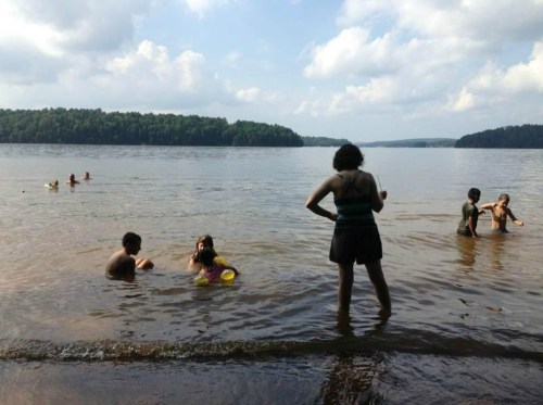 A group of people swimming and playing in a lake on a sunny day, with trees lining the shore in the background.