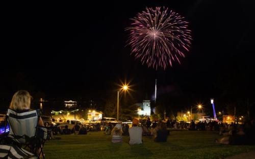 A crowd watches fireworks illuminate the night sky over a small town, with people seated on a grassy area.