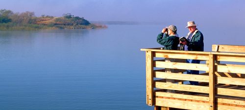 Two people on a wooden dock by a calm lake, one using binoculars and the other reading a book. Misty landscape in the background.