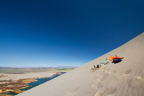 A person in an orange shirt lies on a sandy dune, with a river and green landscape in the background under a clear blue sky.