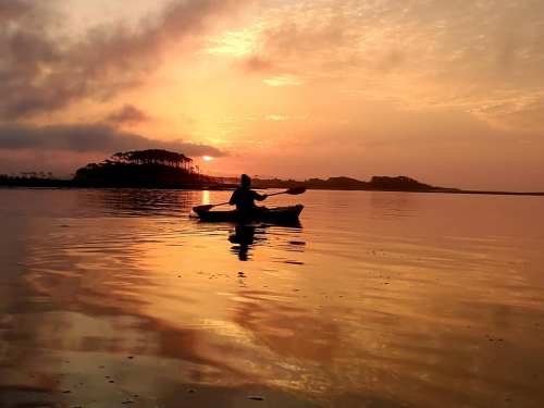 A silhouette of a person kayaking at sunset, with vibrant orange and yellow reflections on the calm water.