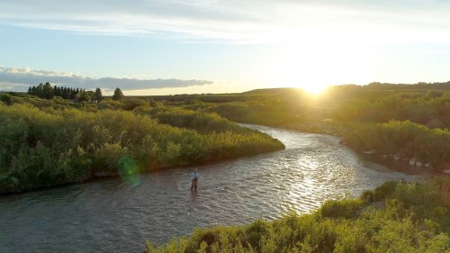 A person stands in a river at sunset, surrounded by lush greenery and a serene landscape.