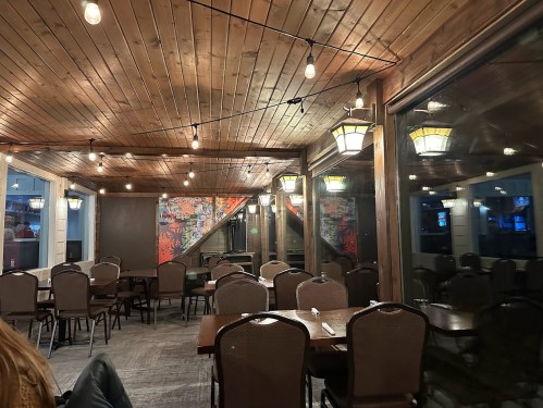 Cozy restaurant interior with wooden ceiling, string lights, and empty tables, featuring a colorful mural on the wall.