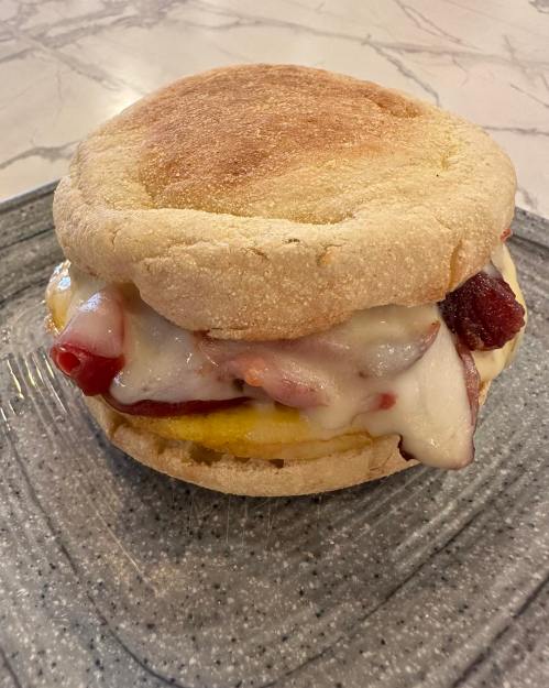 A toasted English muffin sandwich filled with melted cheese, bacon, and ketchup, served on a gray plate.