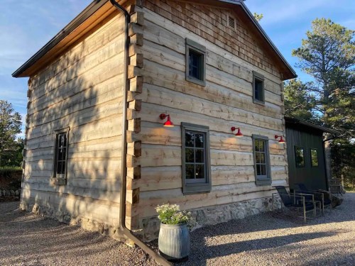 A rustic log cabin with a stone foundation, featuring large windows and outdoor seating in a natural setting.